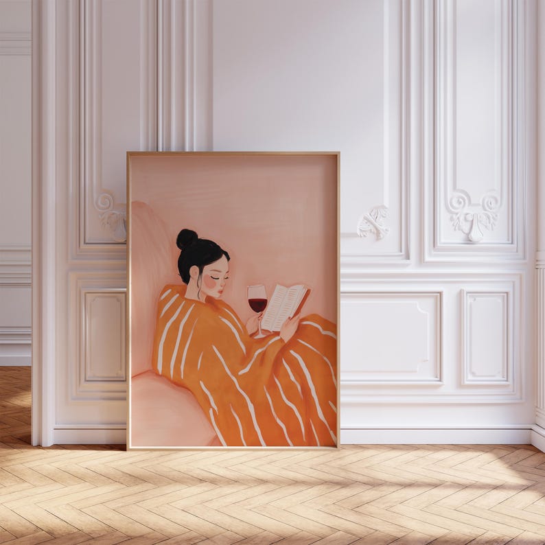 Puede incluir: Una impresi&oacute;n art&iacute;stica enmarcada representa a una persona leyendo un libro y bebiendo vino tinto. La persona est&aacute; envuelta en una manta naranja con rayas blancas. La obra de arte tiene una paleta de colores c&aacute;lidos y est&aacute; ambientada en una habitaci&oacute;n con paneles blancos y suelo de madera.