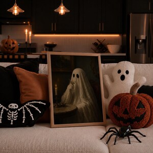 May include: A framed rectangular wall art print featuring a ghost draped in a white sheet sitting at a table with a lit candle and a glass. The artwork is displayed on a couch surrounded by plush Halloween-themed pillows and a plastic spider.