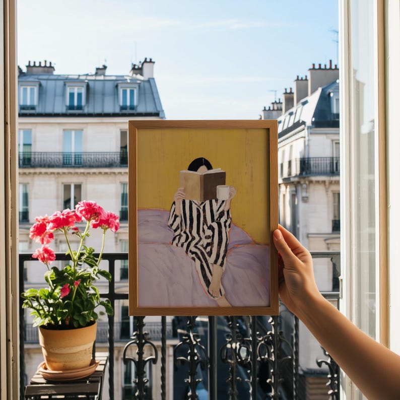 May include: A framed art print depicts a person in striped pajamas reading a book and holding a mug. The artwork has a yellow background and is set against a backdrop of a European cityscape. The frame is made of wood.
