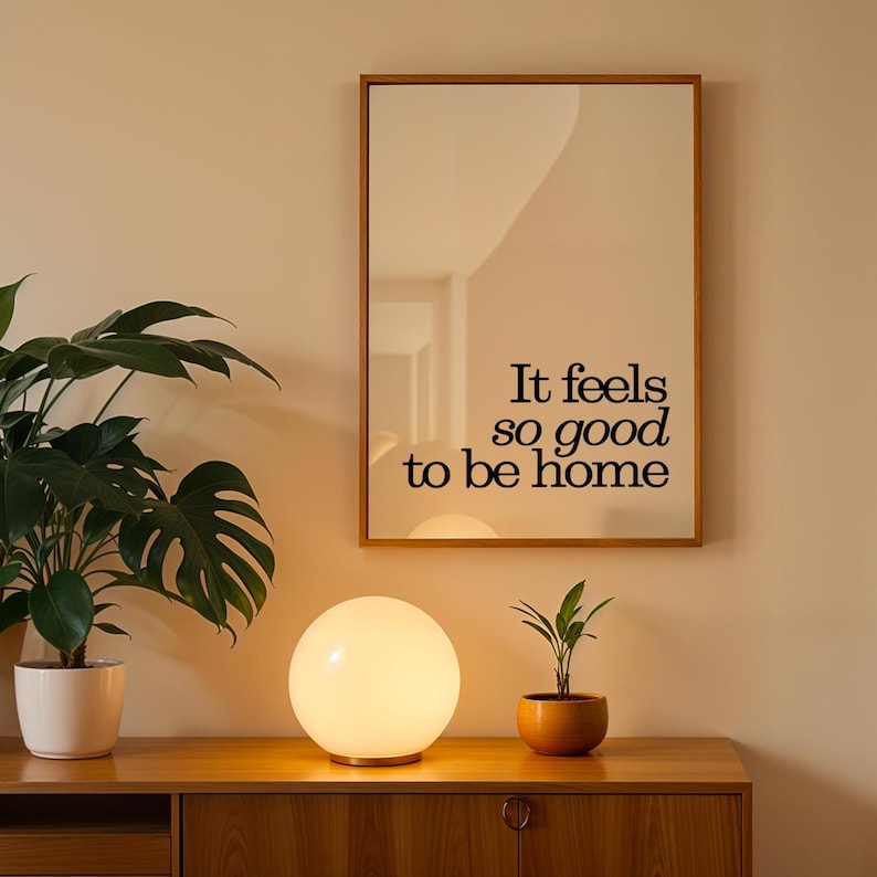 May include: A warm, inviting interior scene featuring a wooden cabinet, a round, glowing table lamp, and a framed mirror with the text "It feels so good to be home." Potted plants add a touch of nature to the decor.