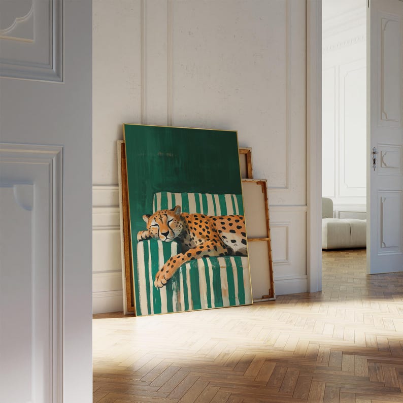 May include: A framed art print featuring a cheetah resting on a green and white striped chair. The cheetah has a tan coat with black spots. The artwork is leaning against a wall in a room with a wooden floor.