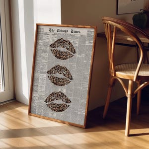 May include: A framed print with three leopard print lips on a vintage newspaper background. The wooden frame displays the text "The Chicago Times". The artwork is leaning against a wall, with a wooden chair visible in the background.