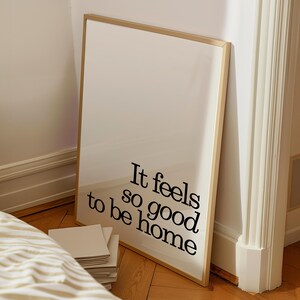 May include: A framed art print with the text "It feels so good to be home" in black lettering. The print has a white background and a thin, gold-colored frame. It leans against a wall on a wooden floor.