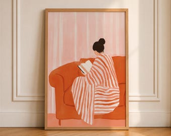 May include: A framed wall art print featuring a person with dark hair in a bun, seen from behind, sitting on a solid orange sofa. The person is wrapped in a white and orange striped blanket while reading an open book. The background is light pink.