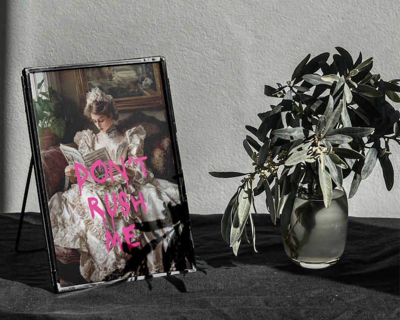 May include: A black framed picture of a woman in a white dress reading a book. The woman is sitting in a chair with a floral pattern. The picture is on a black surface with a vase of green leaves in the background. The text "DON'T RUSH ME" is written in pink on the picture.