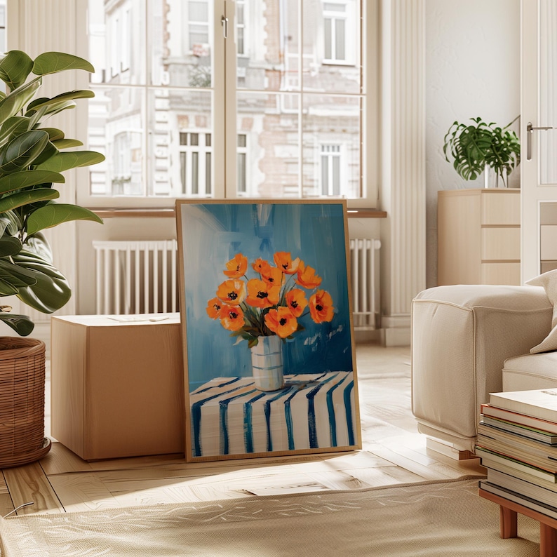 May include: A framed painting depicting orange poppies in a white vase with blue stripes, placed on a blue and white striped table. The background is light blue, with a wooden frame. The painting is in a bright room with a large window.