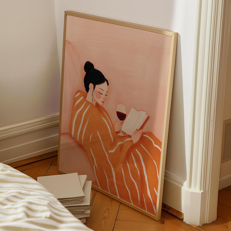 Puede incluir: Una impresi&oacute;n art&iacute;stica enmarcada representa a una persona leyendo un libro y sosteniendo una copa de vino tinto. La persona lleva una bata naranja con rayas blancas. El fondo es rosa suave y el marco es dorado.