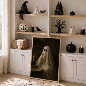 May include: A framed wall art print featuring a ghost draped in a white sheet sitting at a table with a glass, displayed on a white cabinet. The surrounding shelves are decorated with Halloween items including a witch hat, ghosts, pumpkins, and candles.