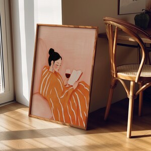 Puede incluir: Una impresi&oacute;n art&iacute;stica enmarcada representa a una persona leyendo un libro y bebiendo vino tinto. La persona lleva una bata naranja con rayas blancas. La obra de arte tiene una paleta de colores c&aacute;lidos y est&aacute; ambientada en una habitaci&oacute;n con una silla de madera.