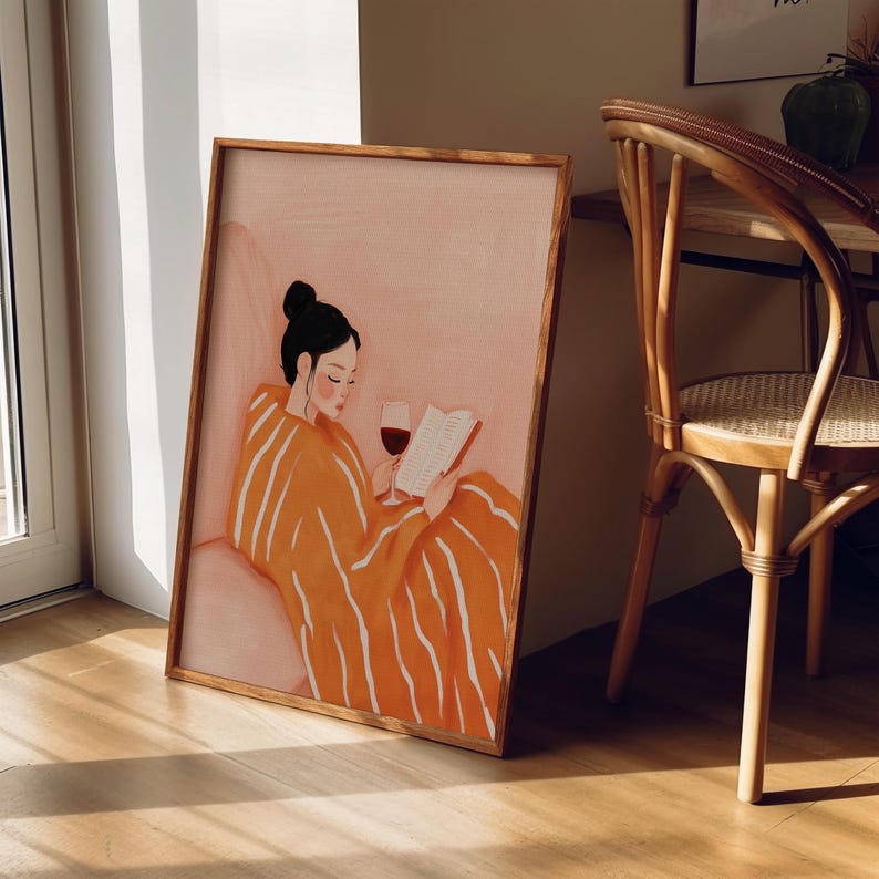 Puede incluir: Una impresi&oacute;n art&iacute;stica enmarcada representa a una persona leyendo un libro y bebiendo vino tinto. La persona lleva una bata naranja con rayas blancas. La obra de arte tiene una paleta de colores c&aacute;lidos y est&aacute; ambientada en una habitaci&oacute;n con una silla de madera.