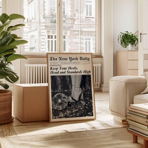 K&ouml;nnte beinhalten: Ein gerahmter Druck der Zeitung "The New York Daily" mit der Schlagzeile "Keep Your Heels, Head and Standards High". Das Schwarz-Wei&szlig;-Bild zeigt eine Discokugel und Beine in High Heels. Der Druck wird in einem hellen Raum ausgestellt.