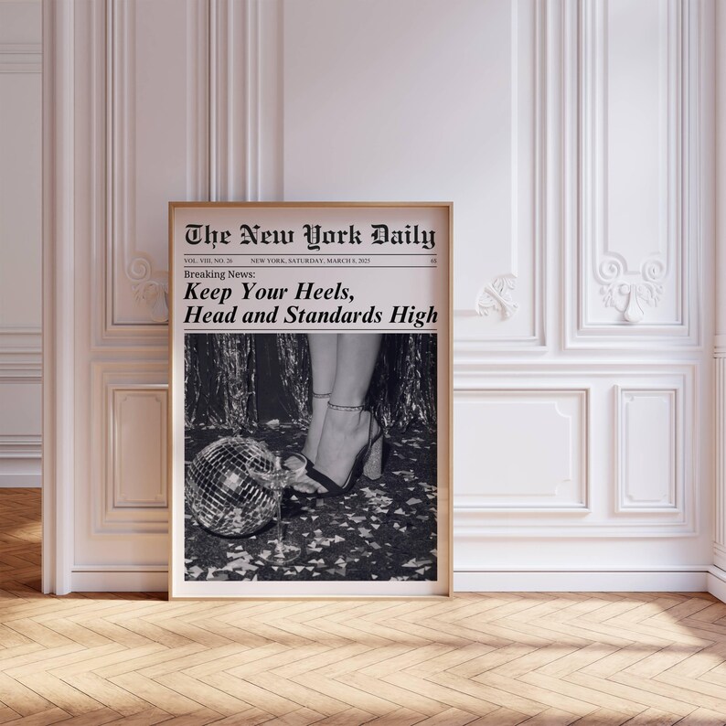 K&ouml;nnte beinhalten: Ein gerahmter Druck einer Zeitungsseite mit dem Titel "The New York Daily". Die Schlagzeile lautet "Keep Your Heels, Head and Standards High". Das Schwarz-Wei&szlig;-Bild zeigt High Heels, eine Discokugel und ein Sektglas.