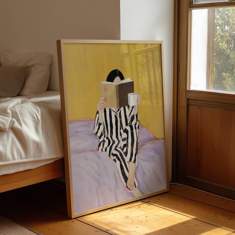 May include: A framed art print depicts a person in black and white striped pajamas reading a book and holding a mug. The artwork is set against a yellow background, with a purple bed and a wooden frame.