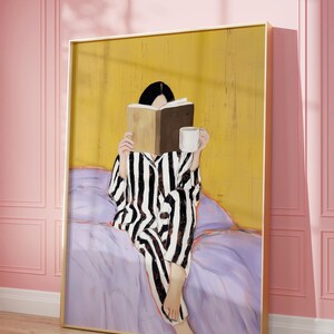 May include: A framed artwork depicts a person in striped pajamas reading a book and holding a mug. The figure is seated on a bed with a purple comforter, against a yellow background. The artwork is in a gold frame, set against a pink wall.