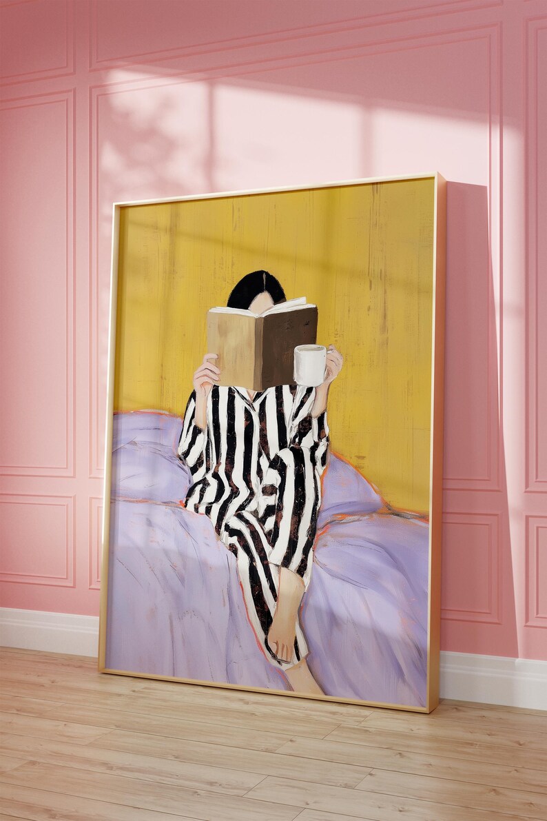 May include: A framed artwork depicts a person in striped pajamas reading a book and holding a mug. The figure is seated on a bed with a purple comforter, against a yellow background. The artwork is in a gold frame, set against a pink wall.