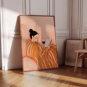 Puede incluir: Una impresi&oacute;n art&iacute;stica enmarcada representa a una persona con cabello negro recogido en un mo&ntilde;o, leyendo un libro y sosteniendo una copa de vino tinto. La persona est&aacute; envuelta en una manta naranja con rayas blancas. Paleta de colores c&aacute;lidos.