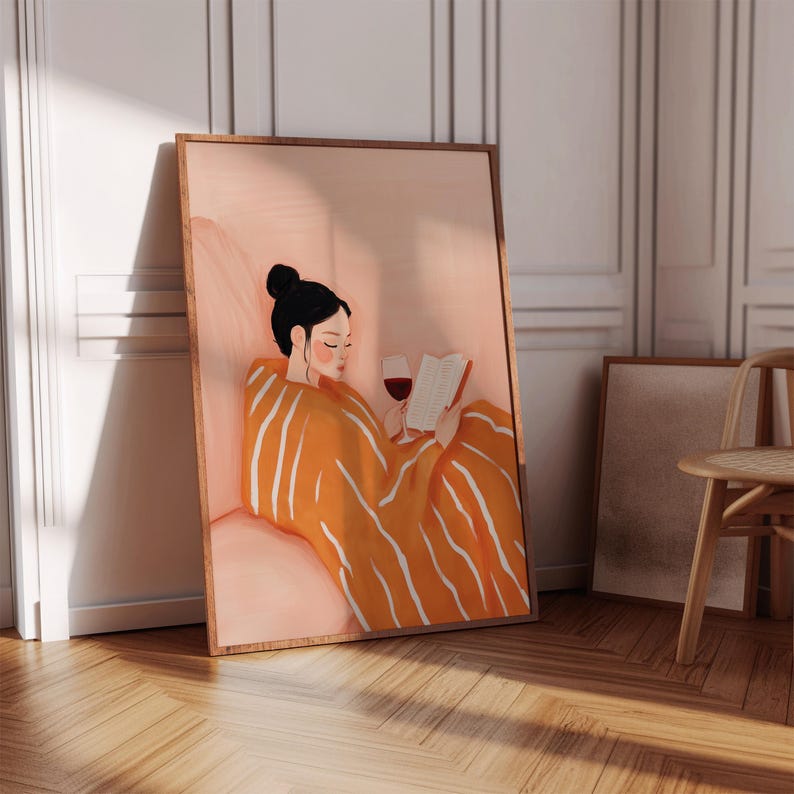 Puede incluir: Una impresi&oacute;n art&iacute;stica enmarcada representa a una persona con cabello negro recogido en un mo&ntilde;o, leyendo un libro y sosteniendo una copa de vino tinto. La persona est&aacute; envuelta en una manta naranja con rayas blancas. Paleta de colores c&aacute;lidos.