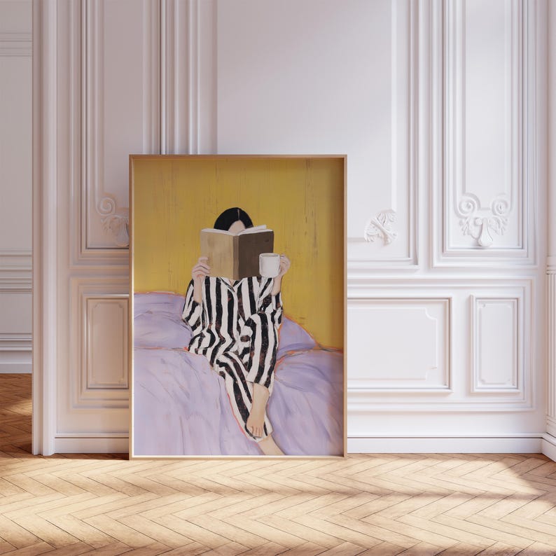May include: A framed art print depicts a person in black and white striped pajamas reading a book and holding a mug. The person is seated on a lavender bed with a yellow background. The artwork is in a gold frame.