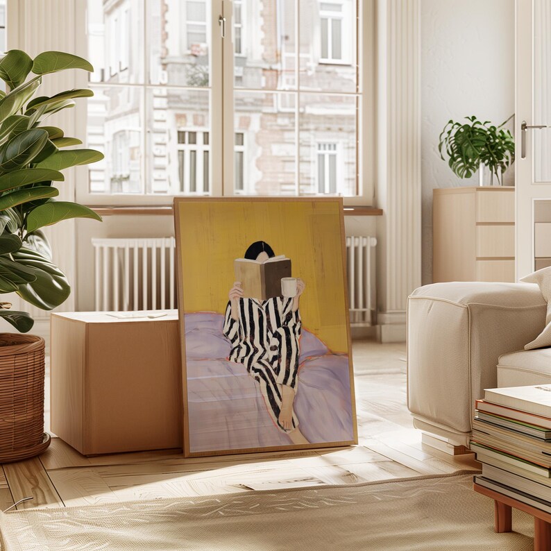 May include: A framed art print depicts a person in black and white striped pajamas reading a book and holding a mug. The artwork has a yellow background and is leaning against a neutral-toned cube. A large plant and a window are in the background.