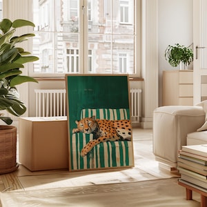 May include: A framed art print featuring a cheetah resting on a striped chair. The artwork has a green background and a wooden frame. The cheetah is orange with black spots. The chair has green and white stripes.