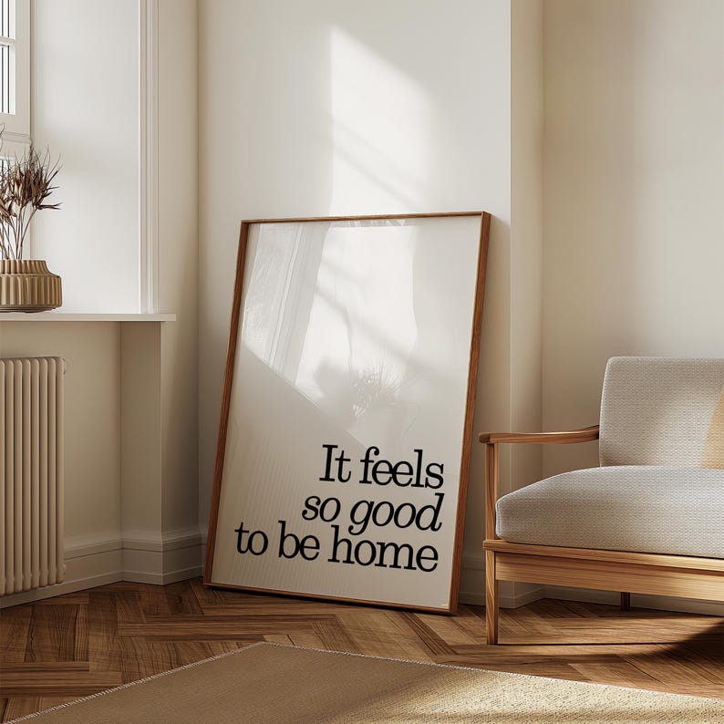 May include: A framed art print with a wooden frame, leaning against a wall. The print features the text "It feels so good to be home" in a black, modern font. The room has a neutral color palette with a beige rug and a wooden armchair.