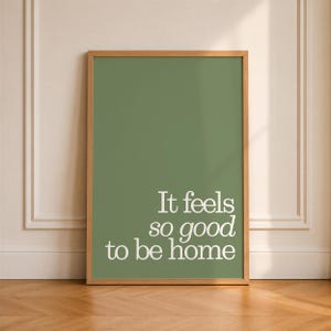 May include: A rectangular wall art print in a light wood frame featuring a solid sage green background with the white text "It feels so good to be home" printed in a clean, serif font in the lower right section of the vertical poster.