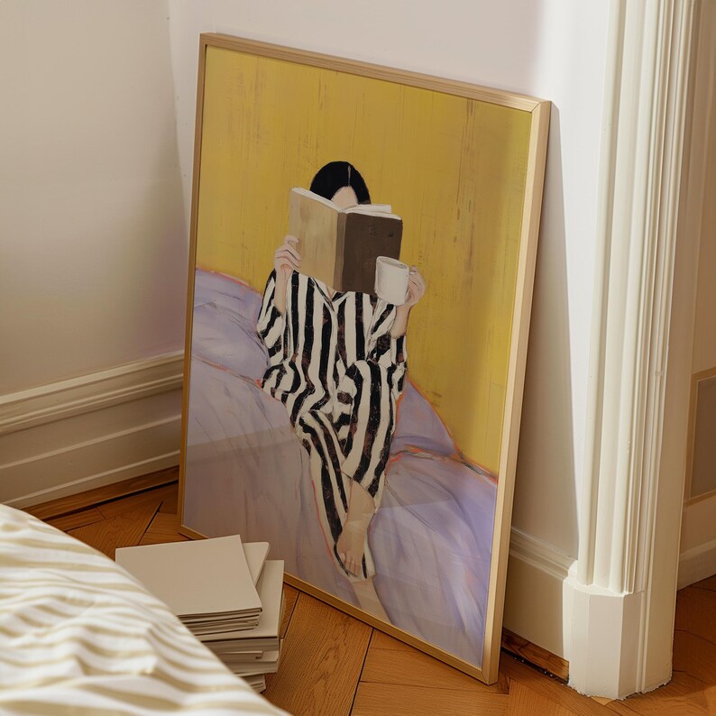 May include: A framed art print depicts a person in black and white striped pajamas reading a book and holding a white mug. The background is yellow, and the person is seated on a purple bed. The artwork has a minimalist style.