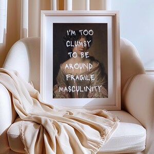 May include: A framed print of a portrait of a woman with a white background. The text "I'm too clumsy to be around fragile masculinity" is written in white paint over the portrait.