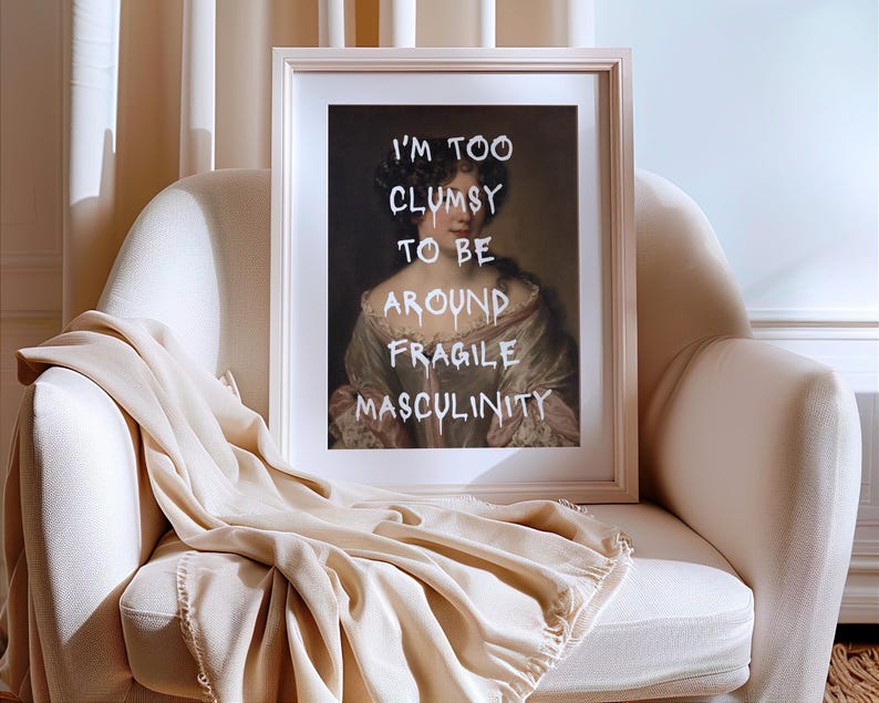 May include: A framed print of a portrait of a woman with a white background. The text "I'm too clumsy to be around fragile masculinity" is written in white paint over the portrait.