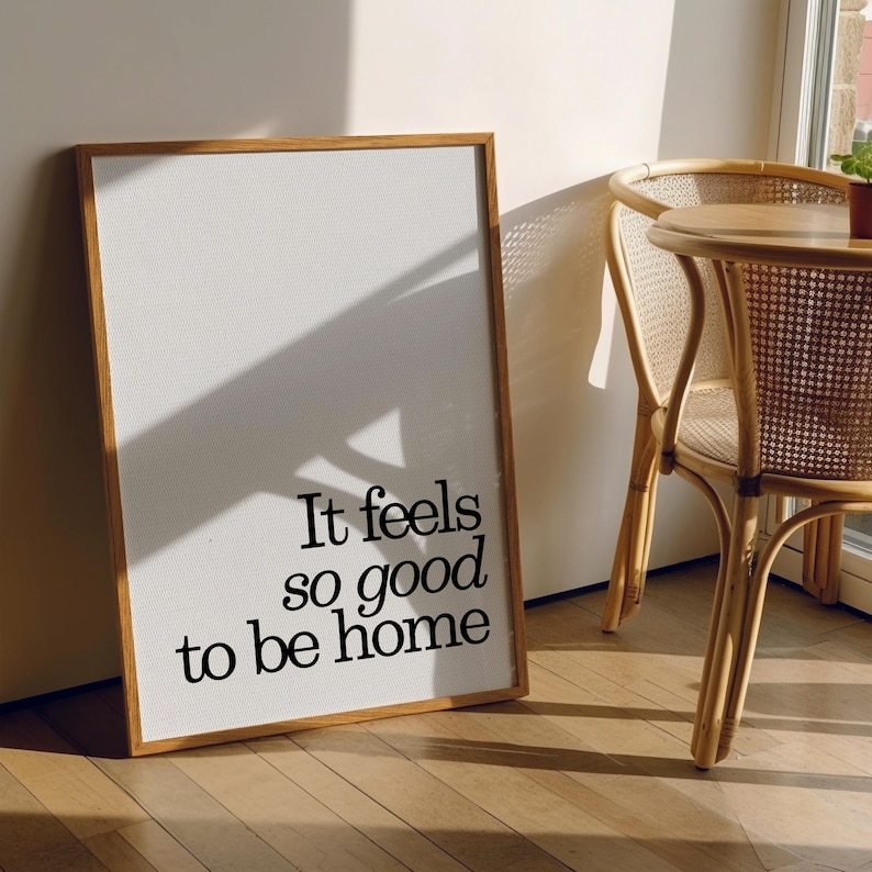 May include: A framed print with the text "It feels so good to be home" in black lettering. The print has a white background and a wooden frame. The image is leaning against a wall next to a wooden chair.