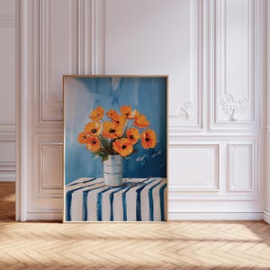 May include: A framed art print depicting orange poppies in a striped vase. The vase is on a blue and white striped table, set against a blue background. The artwork is displayed in a room with white paneling and a wooden floor.