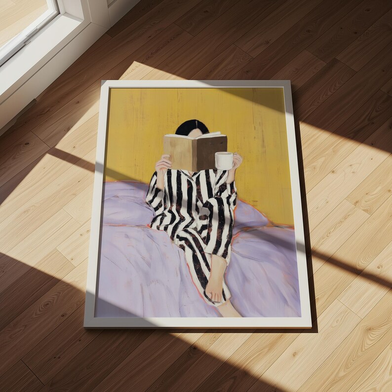 May include: A framed art print depicts a person in black and white striped pajamas reading a book while sitting on a bed. The person holds a white mug. The background is yellow, and the bedspread is lavender.