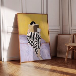 May include: A framed art print depicts a person in striped pajamas reading a book while sitting on a bed. The artwork features a yellow background, a purple bedspread, and a white mug. The frame is a warm brown color, and the overall theme is relaxation and leisure.
