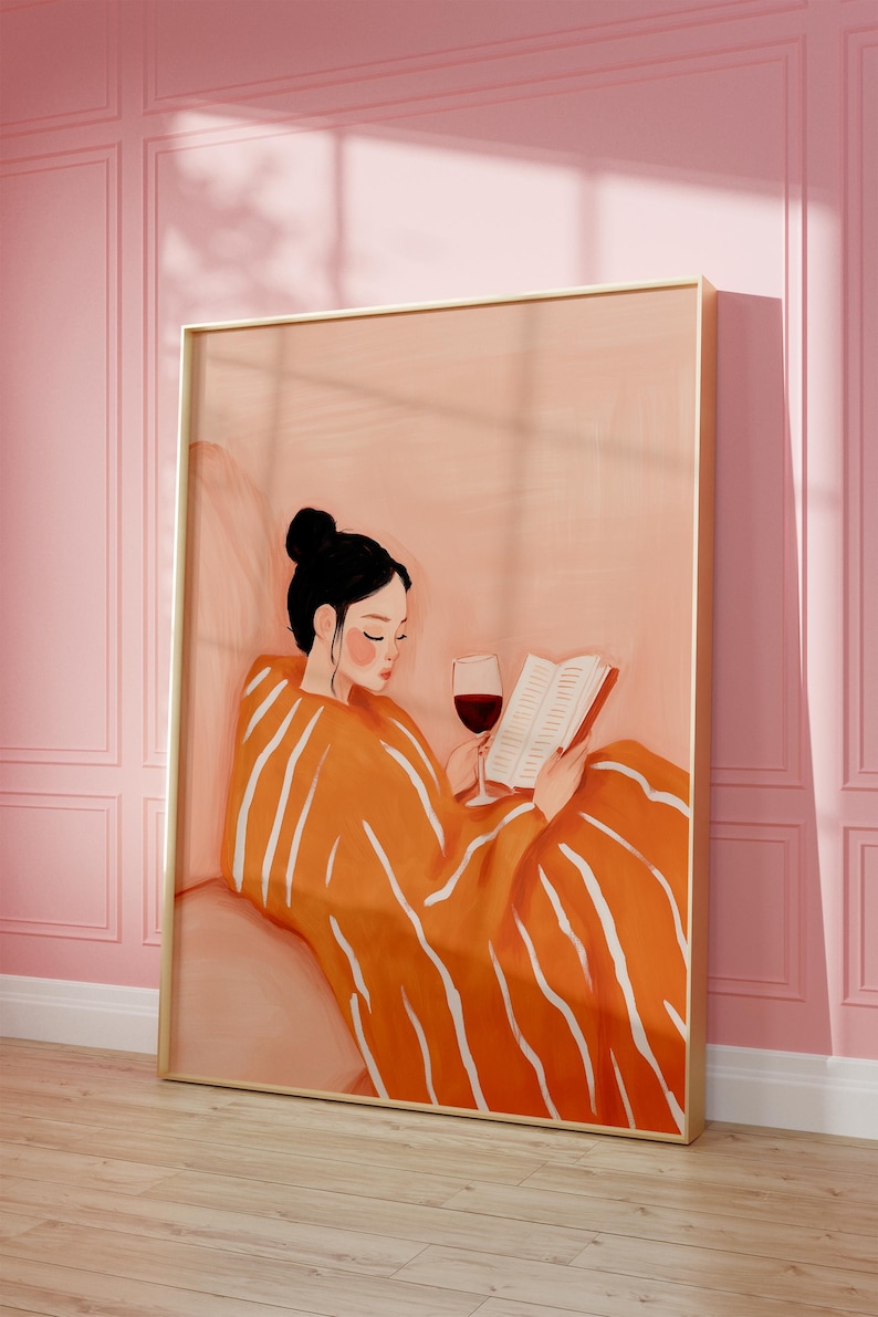 Puede incluir: Una impresi&oacute;n art&iacute;stica que representa a una persona leyendo un libro y bebiendo vino tinto. La persona est&aacute; envuelta en una manta naranja con rayas blancas. La obra de arte est&aacute; enmarcada en un marco dorado y colocada contra una pared rosa.