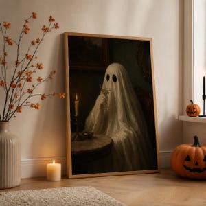 May include: A framed wall art print depicting a ghost draped in a white sheet, sitting at a round table with a lit candle and holding a glass. The ghost has two dark oval eyes. The scene is set in a dark, moody room with a vintage aesthetic.