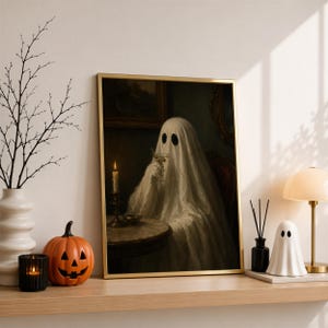 May include: A framed wall art print featuring a ghost draped in a white sheet, sitting at a table with a lit candle and a glass. The ghost has two black oval eyes. The artwork is displayed in a gold frame, capturing a dark, moody atmosphere.