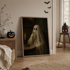May include: A framed wall art print depicting a ghost draped in a white sheet, sitting at a round table with a lit candle and holding a glass. The ghost has two dark oval eyes and is positioned in a dimly lit, moody interior setting.