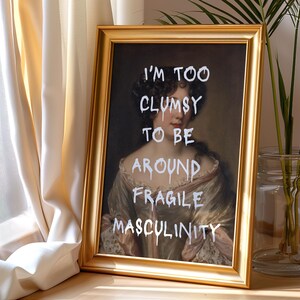 May include: A gold framed print of a portrait of a woman with the text "I'm too clumsy to be around fragile masculinity" written in white paint.