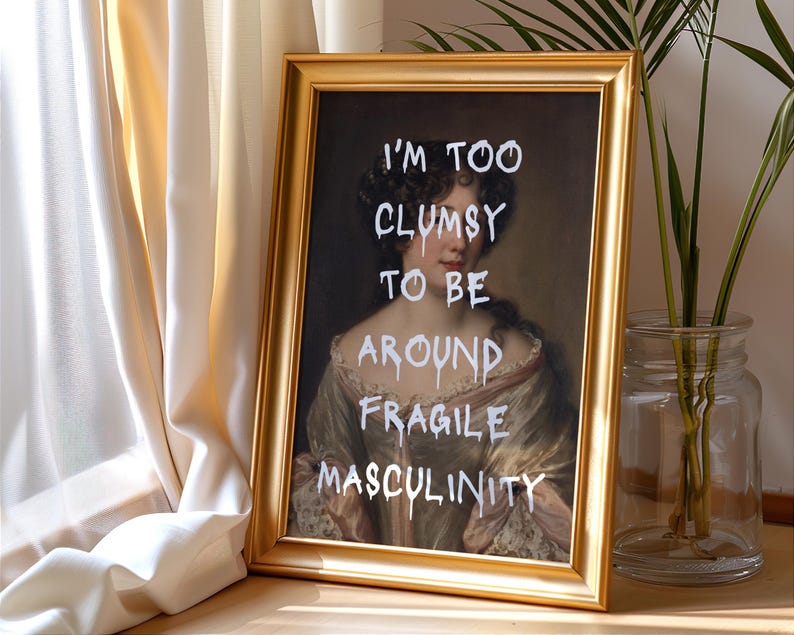 May include: A gold framed print of a portrait of a woman with the text "I'm too clumsy to be around fragile masculinity" written in white paint.