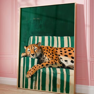 May include: A framed art print featuring a resting cheetah with orange and black spots. The cheetah is lying on a green and white striped cushion against a dark green backdrop. The artwork is in a gold frame and set against a pink wall.