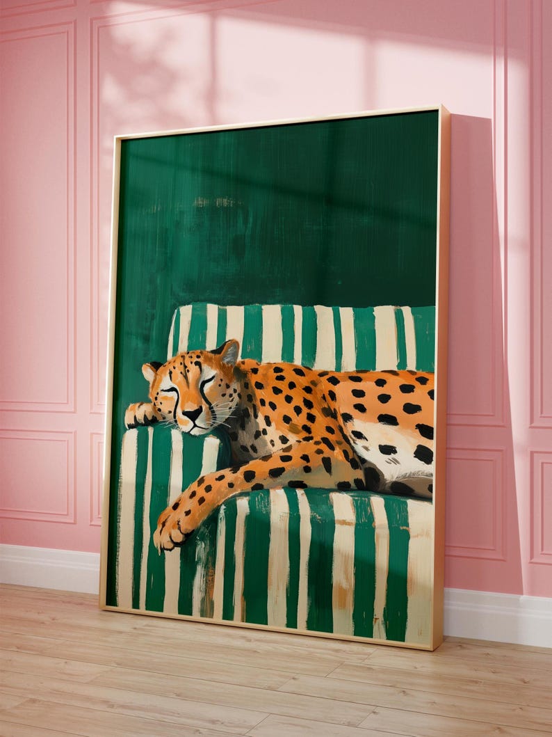 May include: A framed art print featuring a resting cheetah with orange and black spots. The cheetah is lying on a green and white striped cushion against a dark green backdrop. The artwork is in a gold frame and set against a pink wall.