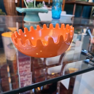 May include: Orange plastic ashtray with a wavy edge. The ashtray is sitting on a glass table.