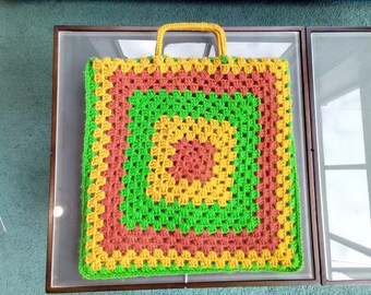 May include: A crocheted tote bag with a green, yellow, and orange geometric pattern. The bag has a handle and is made of yarn.