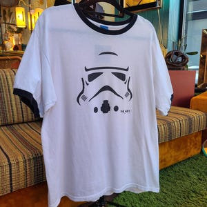 May include: White ringer t-shirt with black trim featuring a black graphic of a Stormtrooper helmet. The text "TK 421" is printed below the helmet. The shirt is hanging on a black hanger.