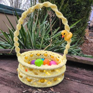May include: A yellow crocheted Easter basket with a yellow rubber ducky on the handle. The basket is filled with colorful plastic Easter eggs.
