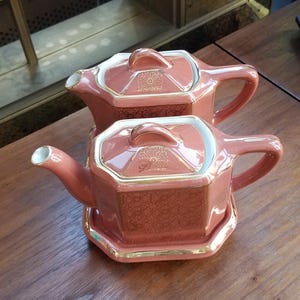 May include: Two pink teapots with gold trim and floral designs. The teapots are on a matching tray.