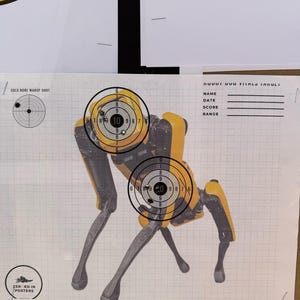 May include: A yellow robot target with two bullseyes. The robot is standing on a gridded white paper with the text "COLD BORE WARUP SHOT" and "ZER-ED IN POSTERS" at the top and bottom of the page. The text "1000T DUO TITALO TARULI" is at the top right corner of the page with the text "NAME", "DATE", "SCORE", and "RANGE" below it.