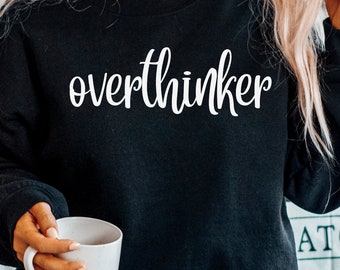 May include: Black sweatshirt with the word "overthinker" in white script. The sweatshirt is a solid black color. The person in the image is holding a white mug.