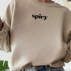 May include: A sand-colored sweatshirt with the word "spicy" printed in black on the front. The sweatshirt is being worn by a person wearing blue denim shorts.