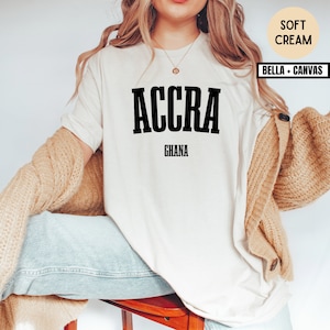 Op de afbeelding: Een zacht cr&egrave;mekleurig T-shirt met het woord "ACCRA" in vette zwarte letters, met "GHANA" eronder gedrukt. Het shirt is van het merk Bella + Canvas. Het model draagt een beige vest en een blauwe jeans.